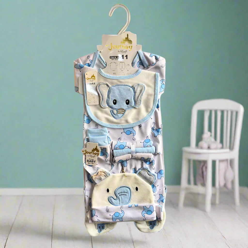 An Alaya Junior Winter Newborn Starter Set - Velvet (11 pieces) featuring a bib, onesie, pants, and hat with adorable elephant and hippo designs hangs on a wooden hanger against a soft teal backdrop. A white chair with plush toys is visible in the background.