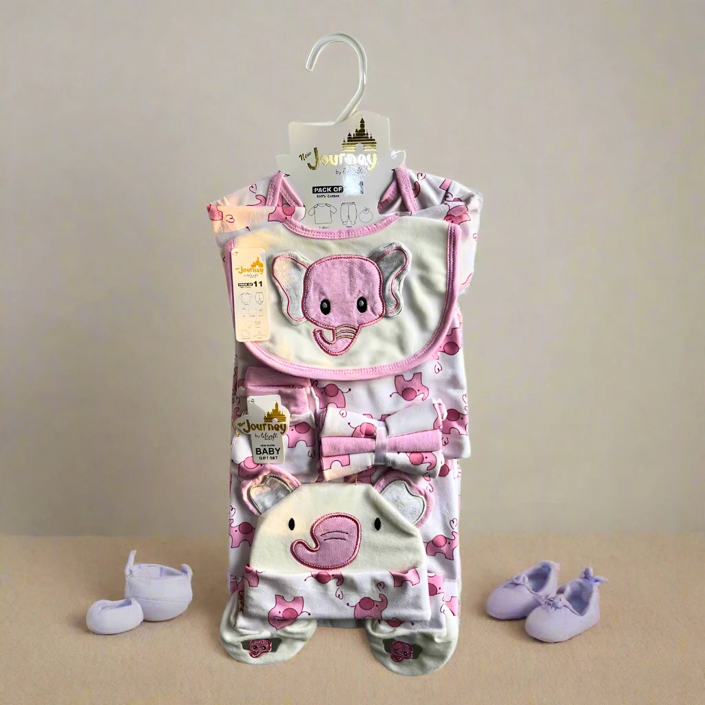 Displayed on a hanger is the Alaya Junior Winter newborn starter set, featuring 11 cozy velvet pieces. Adorned with an adorable elephant theme, it includes a pink and white bib, hat, and onesie for snuggling comfort. Light purple baby booties complete this charming ensemble in the background.