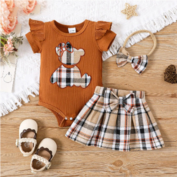 The wooden surface is adorned with the PATPAT Baby Girl Outfit Set: Summer Romper with Plaid Skirt, showcasing a charming cartoon embroidery bear romper paired with a brown onesie, a coordinating plaid skirt, an adorable bow headband, cream shoes accented with brown detailing, accompanied by a lace doily, a small plant, and a starfish decoration.