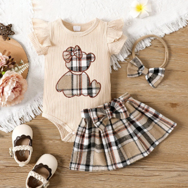 The PATPAT Baby Girl Outfit Set from PATPAT is displayed on a wooden floor, featuring a charming cartoon embroidery bear romper paired with a matching plaid skirt. The ensemble is completed with a plaid bow headband and white shoes highlighted by dark brown detailing, alongside an arrangement of flowers to the side.