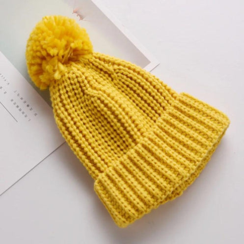 An Alaya Junior 6M-5YRS Kids Winter Hat, featuring a cozy faux fur pom-pom and a folded brim, crafted from stretchable knitted cotton acrylic fabric in yellow, rests on a white surface beside a partially visible open book.