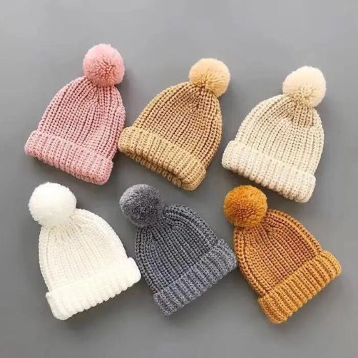 Six Alaya Junior Kids Winter Hats, ideal for winter adventures, are displayed on a gray surface. The colors include pink, beige, cream, white, gray, and mustard yellow. These Baby & Toddler Girls/Boys Faux Fur Pom Pom Beanies are crafted from soft knitted cotton acrylic.