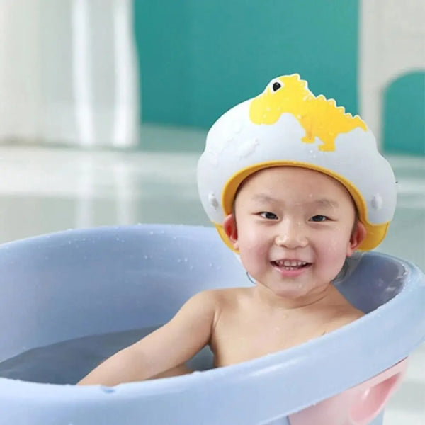 A smiling child enjoys bath time protection in a blue tub, wearing the Adjustable Dinosaur Baby Shower Cap for Kids by Alaya Junior. The cap features an adorable yellow and white dinosaur design, and the background is softly blurred in teal and white tones.