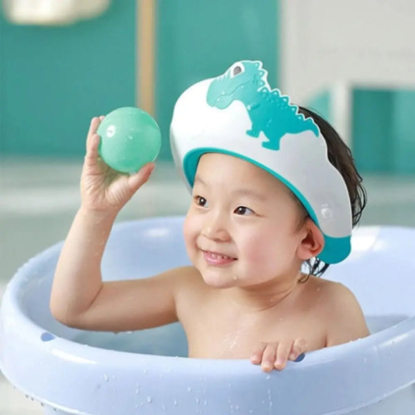 A young child in a bathtub wears the Adjustable Dinosaur Baby Shower Cap for Kids by Alaya Junior, showcasing a charming blue and white dinosaur design that provides bath time protection. Made from child-friendly materials, the child holds a green ball and smiles joyfully, surrounded by a soft teal and white bathroom setting.