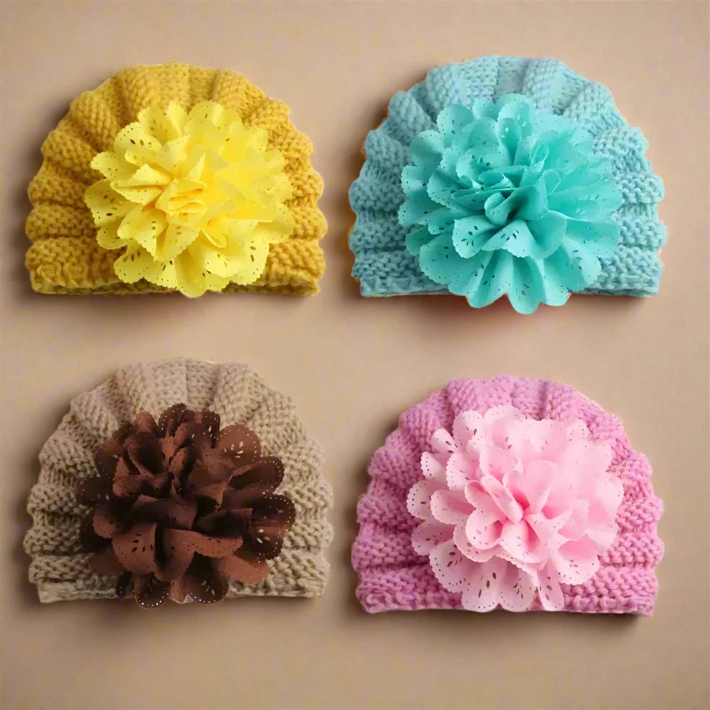 The Alaya Junior Baby Girl Lace Flower Design Knitted Turban Caps, suitable for ages 0 months to 3 years, are displayed on a beige background. These versatile accessories feature large fabric flowers and include options such as yellow with a matching yellow flower, blue with a teal flower, beige adorned with a brown flower, and pink paired with a pink flower.