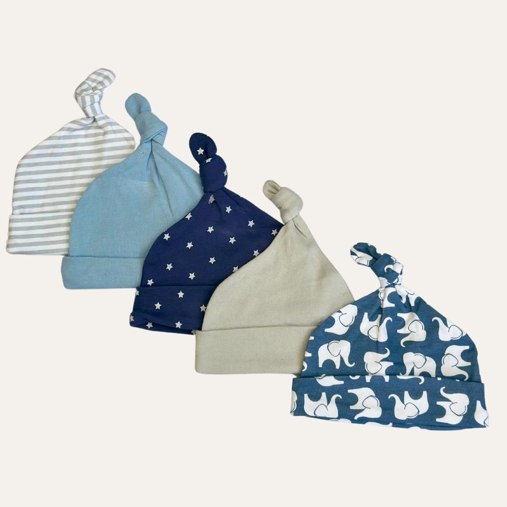Five baby caps with knotted tops are lined up in a row. From left to right, the designs are: gray and white stripes, solid light blue, solid navy with white stars, solid beige, and white elephants on a blue background. The "Baby Blue Dreams: 5-Pack of Infant Boy Cotton Caps" by Alaya Junior is showcased against a white background.