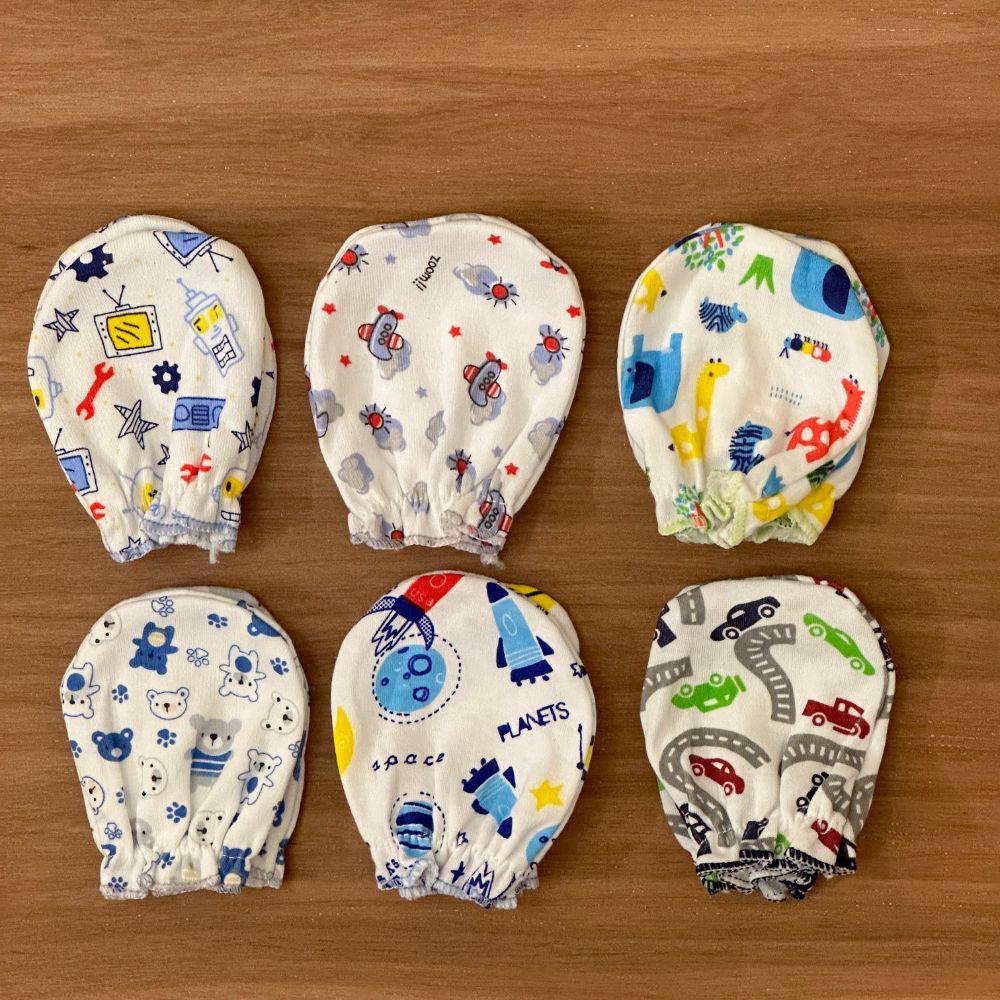 Six Alaya Junior baby boy's mittens (0-6M), featuring vibrant cartoon prints like rockets, dinosaurs, cars, and animals, are laid out on a wooden surface.