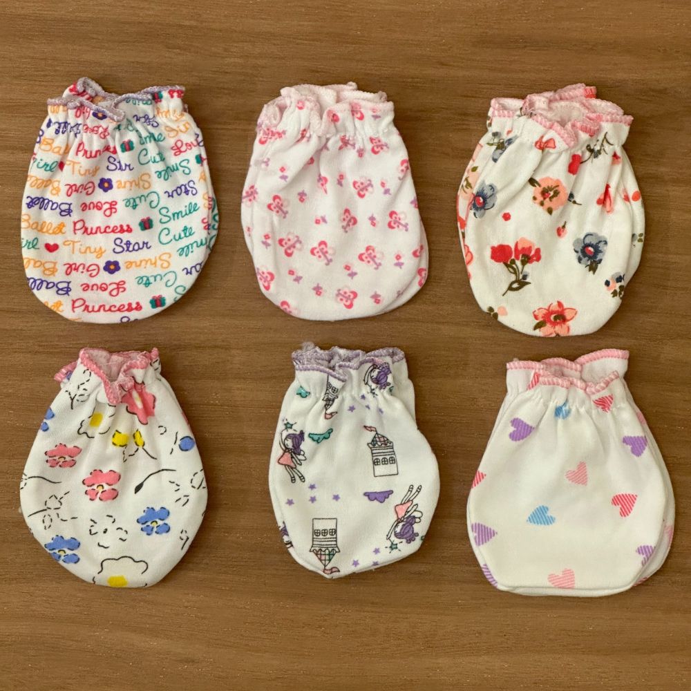 Six pairs of Alaya Junior Baby Girl's 6-Piece Cute Pink Cartoon Mittens (0-6M) are arranged on a wooden surface. Ideal for newborns in Pakistan, these mittens feature designs such as words in colorful fonts, small pink flowers, red and gray flowers, cartoon animals, a whimsical bunny design, and pastel-colored hearts.