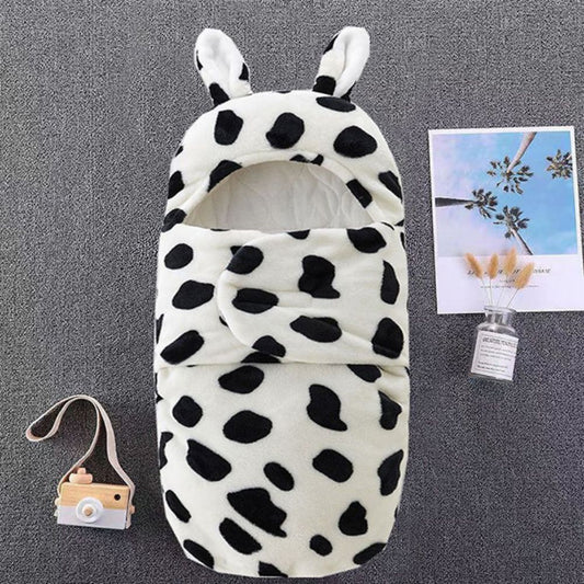 The Alaya Junior Moo Snuggle, an ultra-soft fleece swaddle in a black and white spotted pattern with rabbit ears, rests on gray. Nearby is a wooden toy camera with strap, a floral card, and a clear bottle of dried flowers—ideal for enhancing your baby's whimsical world.