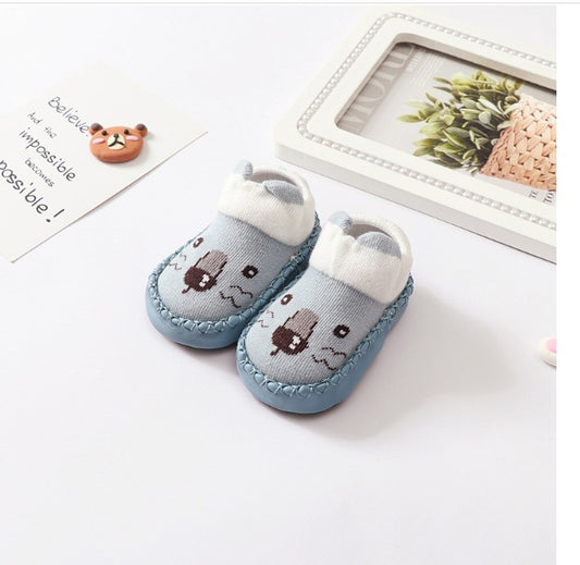 A pair of Whimsical Paws: Winter-Ready Anti-Slip Sock Shoes for Infants by Alaya Junior, featuring blue hues with white cuffs and cartoon bear faces, are placed on a white surface. Nearby are a framed picture of a small plant and a small note with French text and a cartoon design, making them perfect items for those seeking charming newborn baby clothing in Pakistan.
