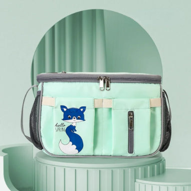 A light green diaper bag and stroller organizer by Alaya Junior, featuring a cartoon fox and the text "hello SPRING" on the front, with a gray zipper. Made of water-repellent material, this diaper bag includes side mesh pockets and two front pouches. It is displayed against a pastel green background with decorative architectural elements.