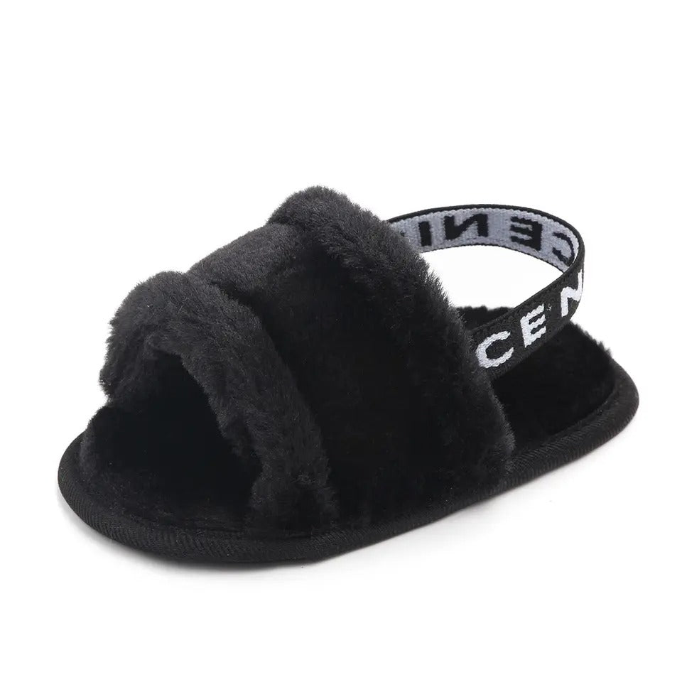The Soft Sole Plush Sandals for Infant Girls by Alaya Junior feature a single black fuzzy slipper with an open-toe design and a thick fur band across the top. This plush sandal includes a back strap with text on it, ensuring a secure fit. The sole is made of solid black material, providing comfort and stability.