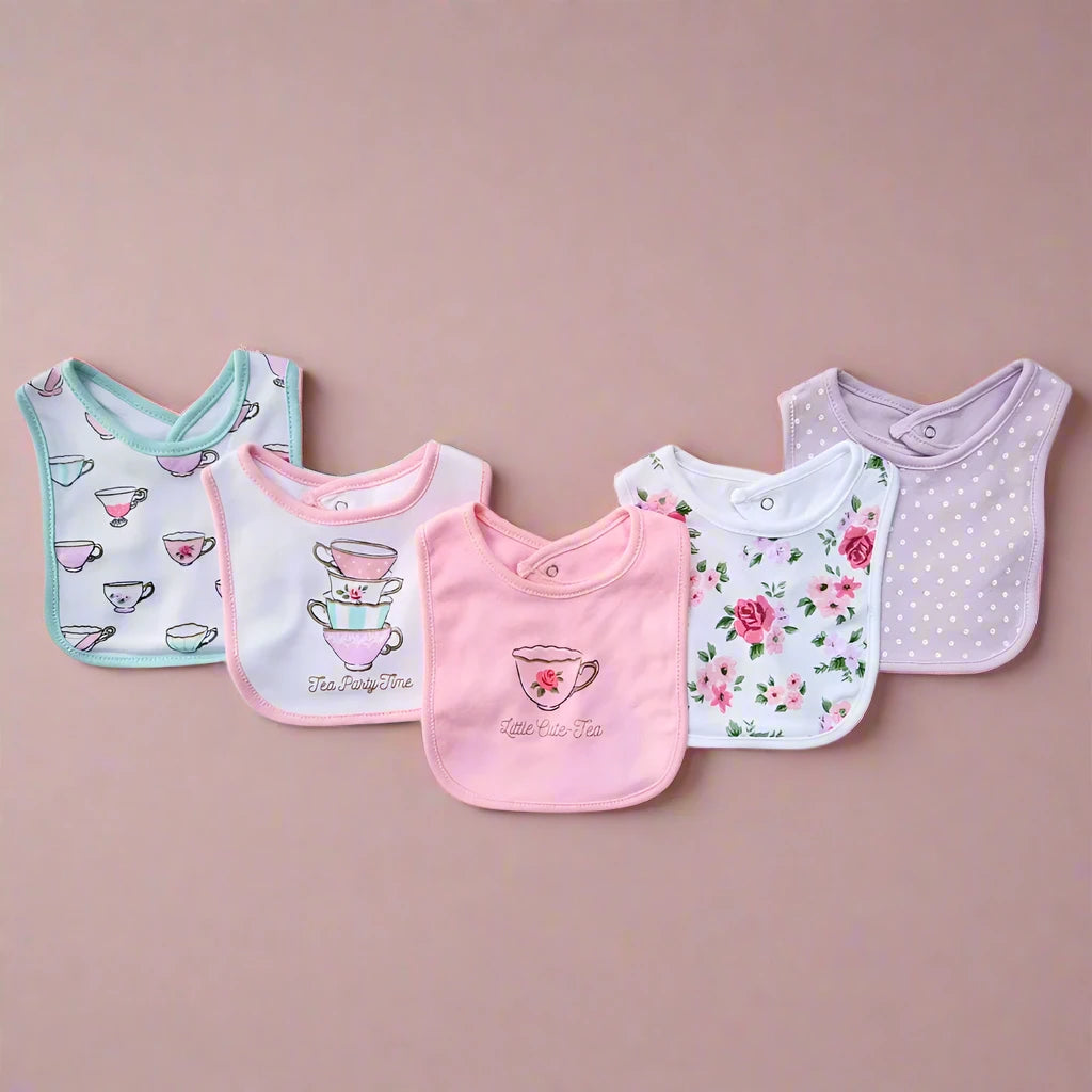 Displayed on a pink background, the Alaya Junior Hudson Baby Infant Girl Cotton Bibs set includes five ultra-soft cotton bibs with unique designs: tea cups, stacked cups, a heart, and floral patterns in pastel pink, white, and lavender shades. Adjustable Velcro straps ensure a snug fit.