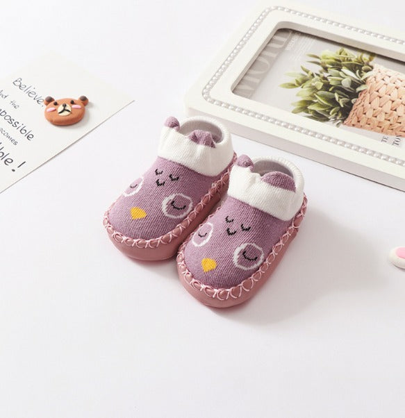 A pair of Whimsical Paws: Winter-Ready Anti-Slip Sock Shoes for Infants from Alaya Junior, featuring pink hues, cute owl designs with closed eyes and yellow beaks, rests on a white surface. Surrounded by a framed picture of a plant and an illustrated bear on paper, these adorable slippers are perfect for adding charm to your newborn baby's wardrobe in Pakistan.
