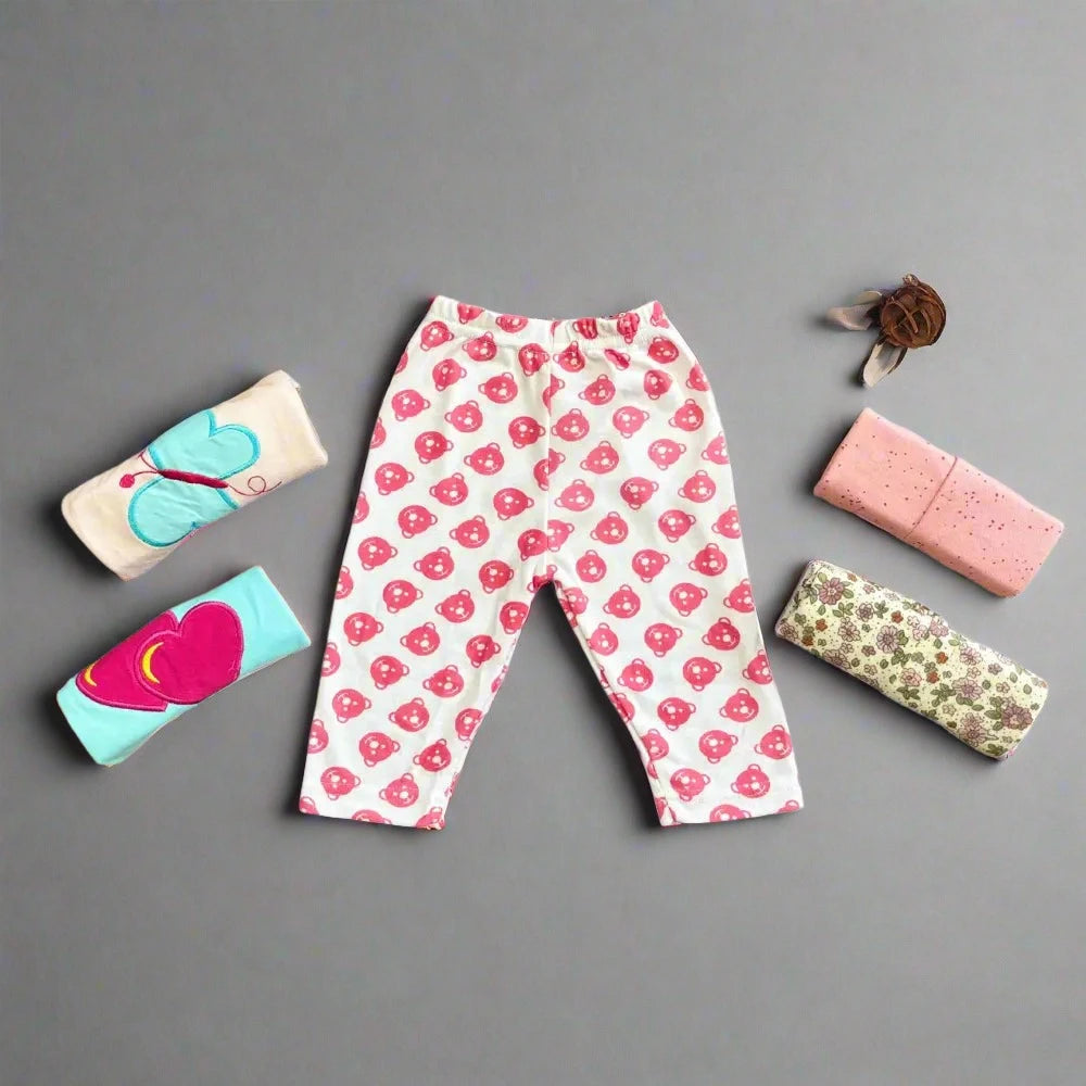 A pair of trousers from the "Pack of 5 Baby Girl Trousers Soft Fabric" by Alaya Junior, featuring a pink and white heart pattern, is laid out on a gray background. Surrounding the pants are four neatly rolled-up shirts in various pastel colors and designs, along with a brown headband adorned with a flower detailβideal for newborn baby clothing in Pakistan.