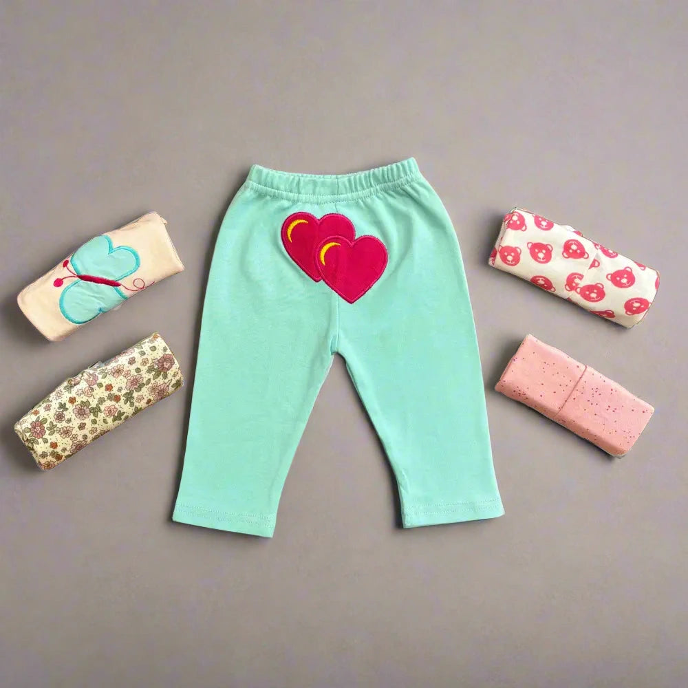 A set of light blue Alaya Junior trousers, featuring heart-shaped patches on the back, is laid flat against a gray background. Surrounding the trousers are five neatly rolled-up baby clothes in various colors and patterns, including florals, strawberries, and polka dotsโan ideal collection for newborn and infant clothing in Pakistan.