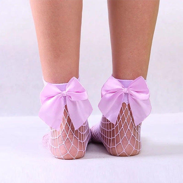 A person wearing Alaya Junior's Big Bow Mesh Socks for Baby Girls, featuring pink bows on the back. The socks and bows pop against a pale background, showcasing the person's lower legs and feet from behind. The image exudes a minimalistic, stylish aesthetic reminiscent of infant clothing designs from Pakistan.