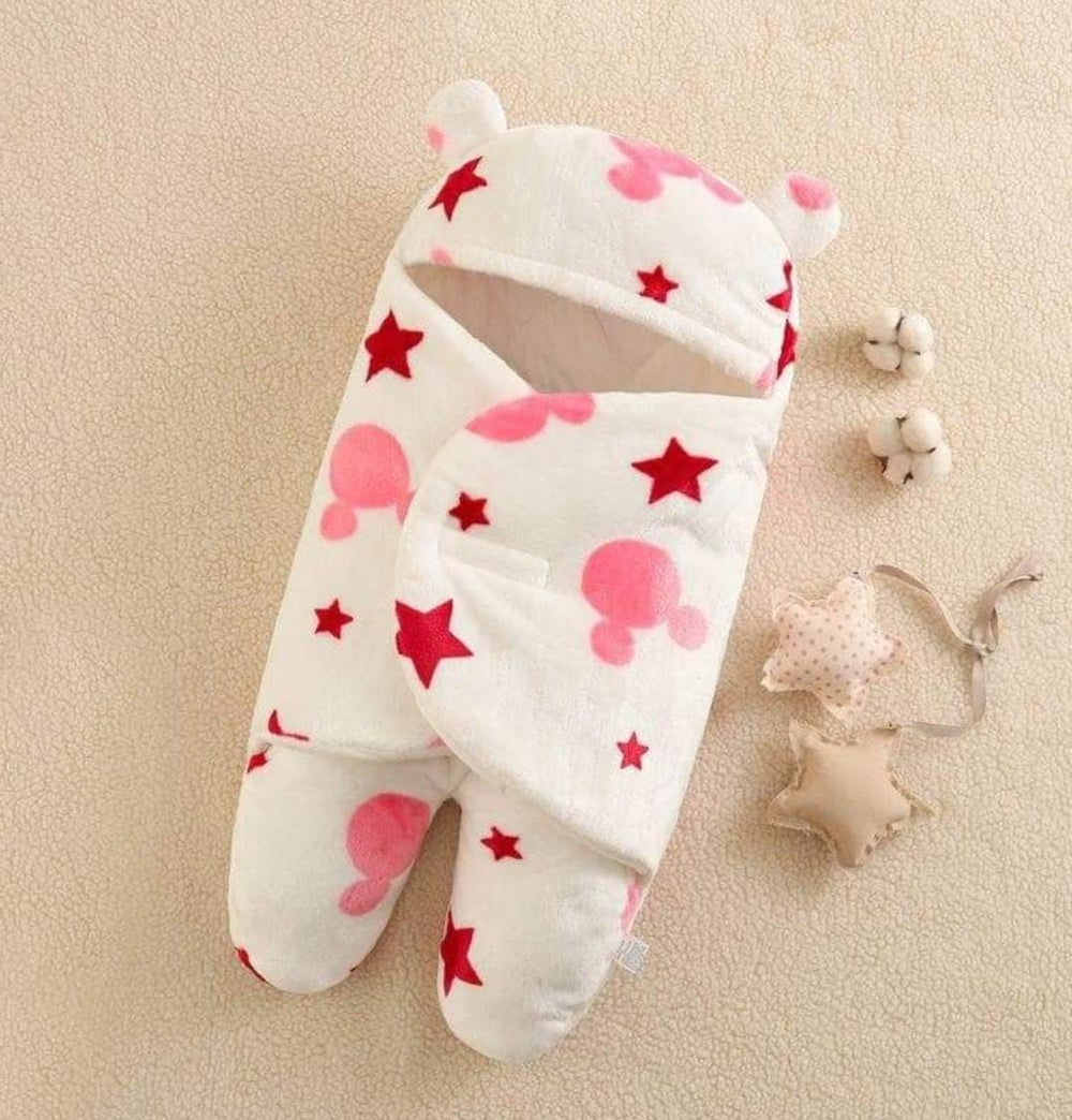The Cozy Coral Embrace: Multifunctional Baby Fleece Swaddle Blanket by Alaya Junior features a Mickey design on a white background adorned with red stars and pink patterns. Made from soft coral fleece, this all-season comfort piece includes bear ears on the hood and is showcased on a beige surface next to two cotton flowers and star-shaped toys.