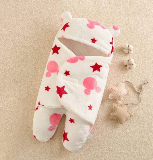 The Cozy Coral Embrace: Multifunctional Baby Fleece Swaddle Blanket by Alaya Junior features a Mickey design on a white background adorned with red stars and pink patterns. Made from soft coral fleece, this all-season comfort piece includes bear ears on the hood and is showcased on a beige surface next to two cotton flowers and star-shaped toys.