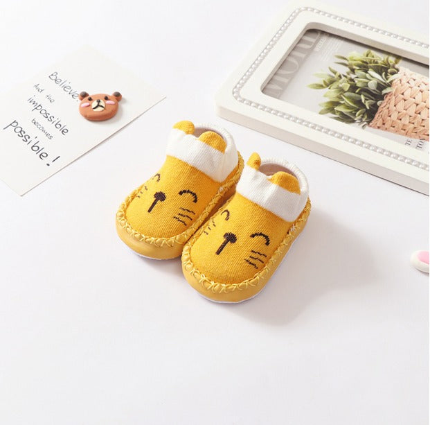 A pair of Whimsical Paws: Winter-Ready Anti-Slip Sock Shoes for Infants from Alaya Junior, featuring a cute yellow cat face design with white trim. The shoes are placed on a white surface next to a card that reads "Believe the impossible is possible" and a framed plant picture. A small bear figurine is also visible, perfect for newborns in Pakistan's finest infant clothing collections.