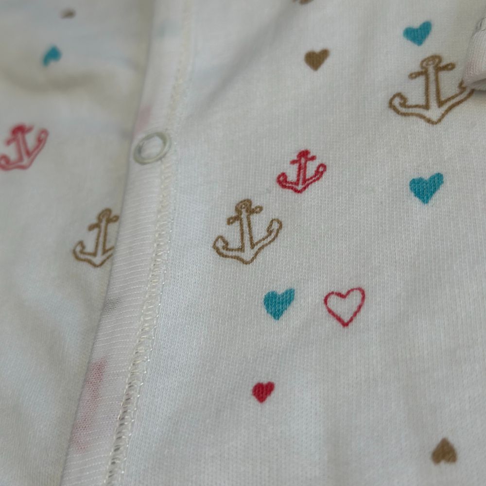 Close-up of a white fabric with small prints of anchors and hearts in vibrant red, blue, and brown colors. This fabric is part of the Juniors Duo Footed Rompers Set (Skin and White) by Alaya Junior. It features a central seam and a metal snap button, indicating it is an adorable piece of baby clothing from Pakistan, similar to a charming newborn onesie.