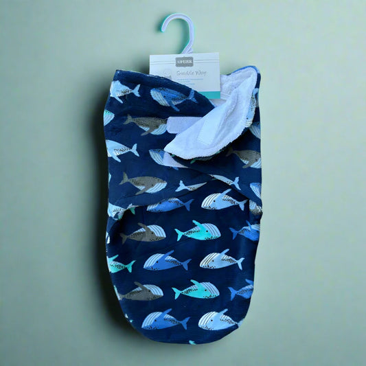 An Alaya Junior Cozy Winter Swaddle with a blue fish design hangs on a hook against a light green background. This baby wrap, featuring its soft white interior and a label at the top, promises to keep your little one snug and warm.