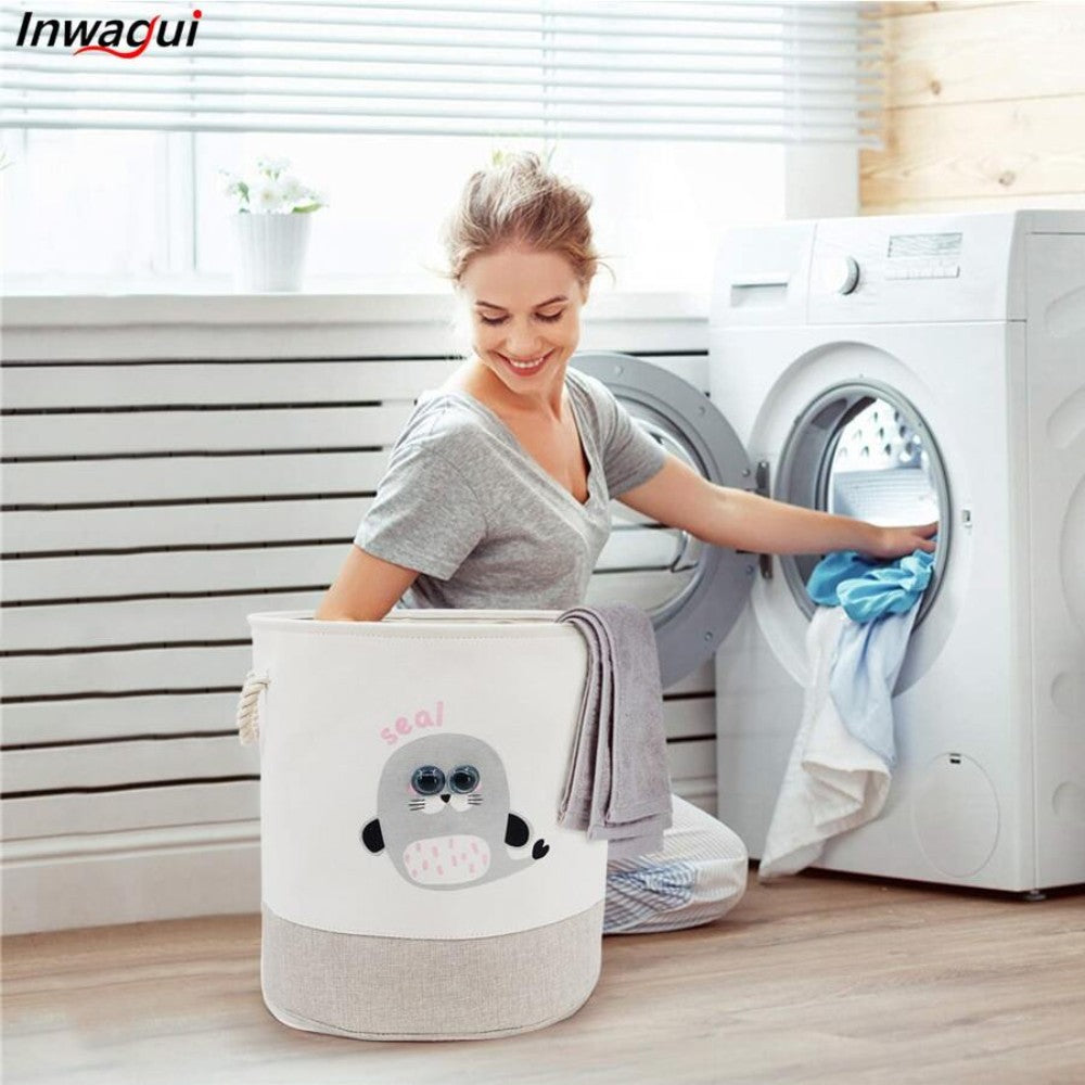 A person smiles as they kneel in front of a washing machine, adding laundry. Nearby, there's a SHEIN Kids Large Folding Laundry Basket featuring an adorable seal design. The room is bright and airy with striped walls and plants on the windowsill.