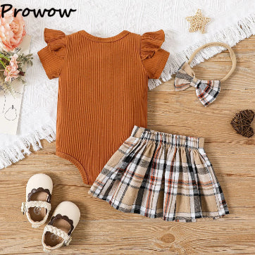 The PATPAT Baby Girl Outfit Set includes a rust-colored ribbed romper with ruffled sleeves, combined with a plaid skirt. Nearby, a coordinating plaid headband, white baby shoes, and a pine cone are displayed on a lace-trimmed surface alongside a bouquet of flowers and starfish.