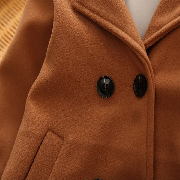 Close-up image of the PAT PAT Toddler Girl/Boy Lapel Collar Double Breasted Coat by Alaya Junior, showcasing two black buttons and a notched lapel. The brown coat's fabric appears to be wool or a similar material, with part of a pocket visible on the left side. It is lying against a textured background reminiscent of delicate designs in baby clothing from Pakistan.