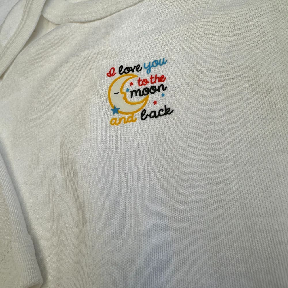 This image showcases a Carter's Bliss bodysuit from Alaya Junior, made of organic cotton in a pristine white shade. The front features vibrant embroidery of a smiling crescent moon nestled among stars, with the heartfelt message "I love you to the moon and back" displayed in red, blue, and black letters. These bodysuits offer timeless style with whimsical accents for those who appreciate both elegance and fun.