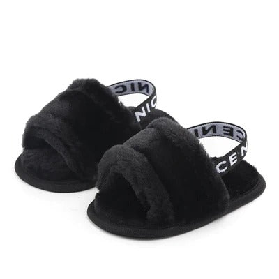 The Alaya Junior Soft Sole Plush Sandals for Infant Girls are designed with black furry slip-on uppers, open toes, and elastic back straps adorned with the word "NICE" printed in white. These cozy and stylish sandals feature a furry texture that covers both the upper part and the sole.
