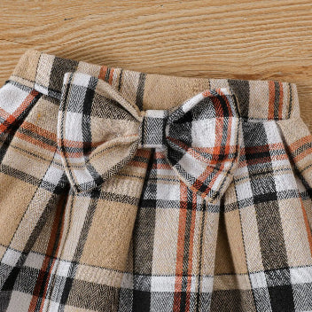 A close-up shot of the PATPAT Baby Girl Outfit Set captures the adorable plaid skirt with a bow on top. This charming pleated skirt features a pattern of tan, white, black, and orange stripes and is displayed on a wooden surface. It's perfect for pairing with a cartoon embroidery bear romper from PATPAT.