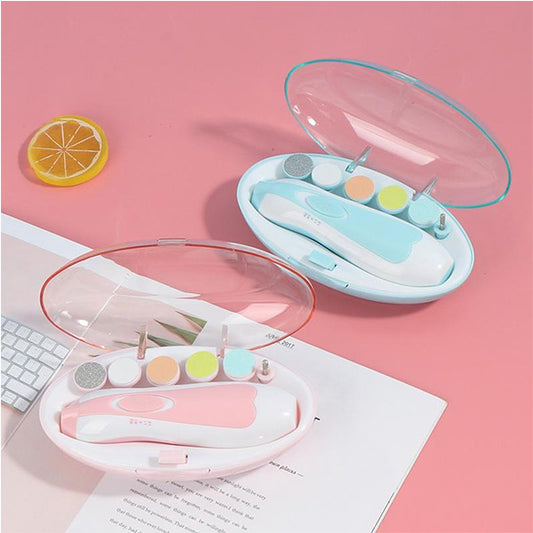 Two Electric Baby Nail Trimmers with 6 Grinding Heads by Alaya Junior are displayed on a pink surface in open cases. One kit features a blue case, and the other has a pink case. Each trimmer comes with various attachments, including buffing heads and files. A keyboard, a lemon slice, and some baby clothing from Pakistan are also visible.