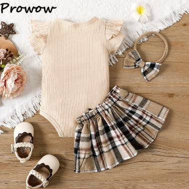 The PATPAT Baby Girl Outfit Set: Summer Romper with Plaid Skirt is an adorable ensemble featuring a beige ribbed onesie with frilled sleeves, beautifully paired with a plaid skirt and headband. Complementing the outfit are cream shoes adorned with brown trim, all stylishly arranged on a wooden surface surrounded by flowers and a lace doily for a picture-perfect look.
