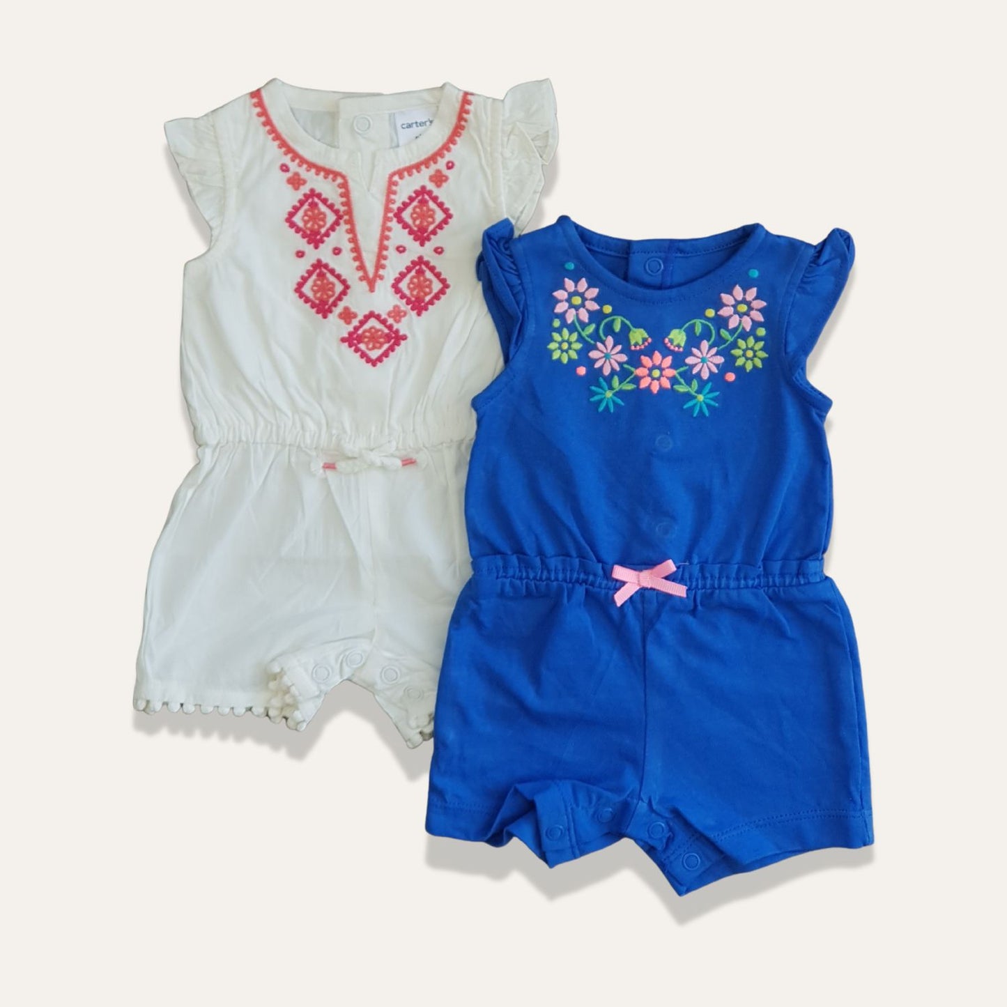 Two baby rompers are displayed side by side. The one on the left, from the Alaya Junior brand, is white with ruffled sleeves, a pink embroidered pattern, and snaps at the bottom. The romper on the right is blue with pink, green, and white floral embroidery and features a pink bow at the waist—both perfect examples of stylish infant clothing in Pakistan from Alaya Junior's "Baby Girls Embroidered Jumpsuit-Thailand" collection.