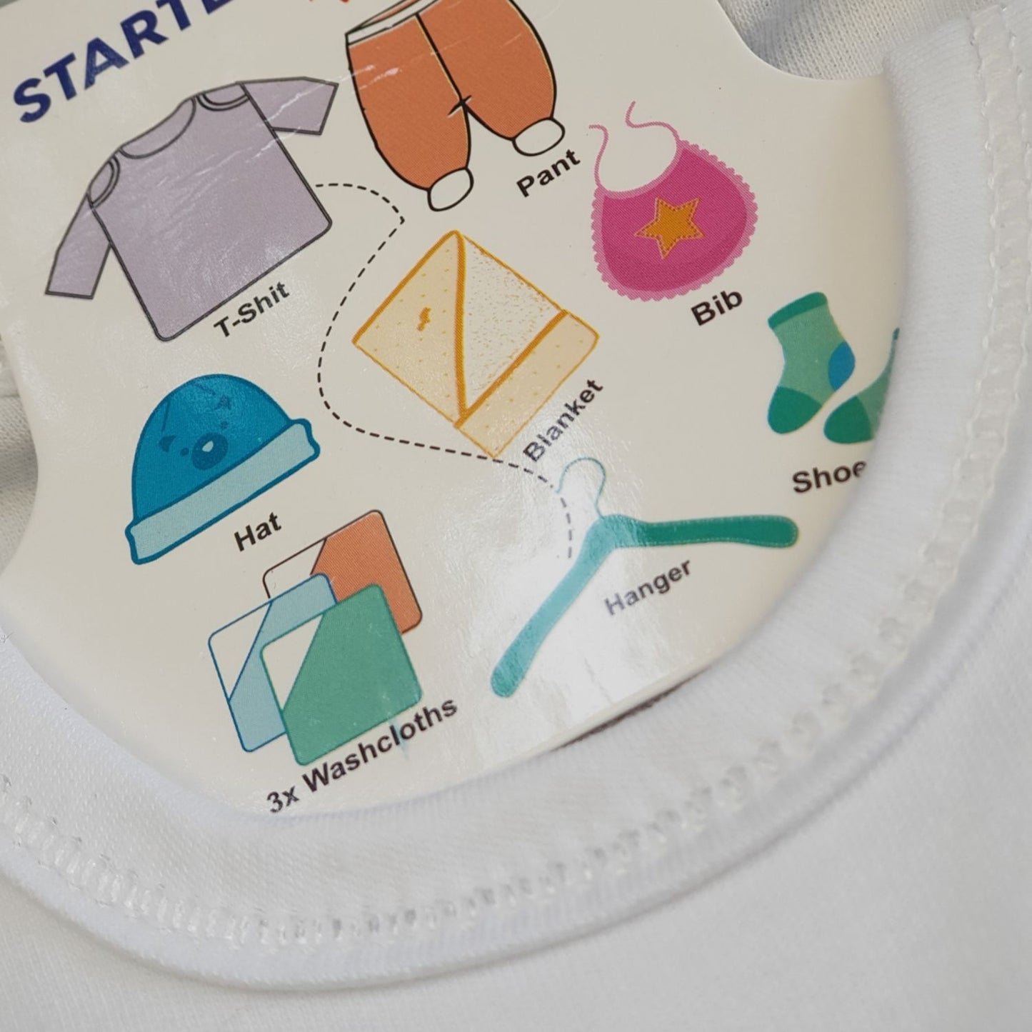 A clothing tag with colorful illustrations of newborn baby items, including a T-shirt, pants, bib, hat, blanket, washcloths, hanger, socks, and shoes from the Newborn Essentials 10-Piece Starter Pack by Alaya Junior. Sourced from the finest infant clothing in Pakistan, the tag is attached to a piece of white fabric.
