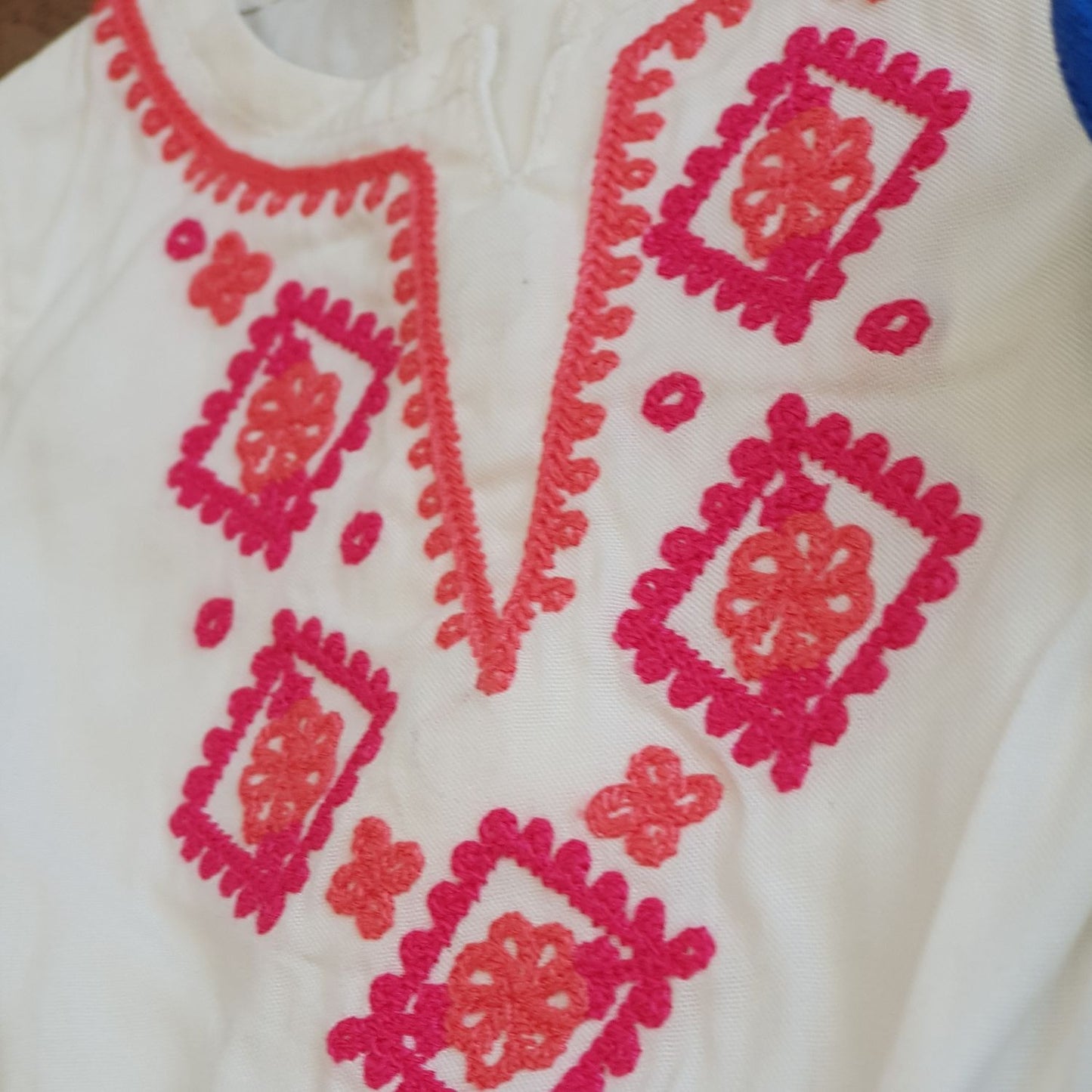 A close-up of the Baby Girls Embroidered Jumpsuit-Thailand by Alaya Junior showcases a white fabric with intricately embroidered patterns, ideal for newborn baby clothing in Pakistan. The embroidery boasts red and pink flowers and geometric shapes such as squares and diamond-like patterns, surrounding a central vertical opening adorned with detailed red stitching.