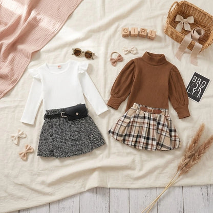 Children's clothing set with a white shirt, brown sweater, and plaid skirt on a neutral background.