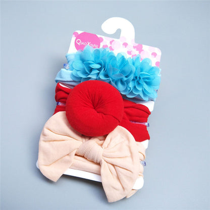 The Alaya Junior Baby Girls' Adorable Trio Three-Piece Headband Set, ideal for baby girl fashion, is neatly displayed on a white hanger. This stylish collection of hair accessories includes a headband with a turquoise floral design, one with a red donut shape, and another featuring a beige bow. The packaging is adorned with a pink label showcasing a heart pattern.