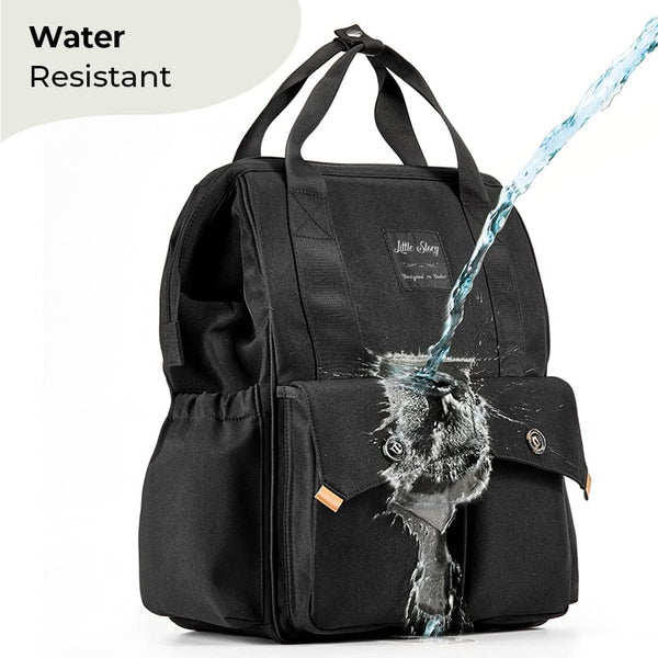 The Ellie & Luke Multifunctional Diaper Bag in black showcases its water-resistant feature and sleek design with water splashing over it. It includes two front pockets with button closures, sturdy top handles, and insulated bottle holders for convenience.