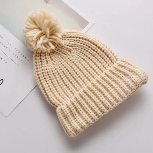 An Alaya Junior 6M-5YRS Kids Winter Hat in a cozy cream color, featuring a large faux fur pom-pom on top, is placed on a flat surface next to a partially visible magazine, ready for winter adventures. This beanie includes a wide ribbed brim, crafted from soft knitted cotton acrylic fabric that ensures the perfect fit for baby and toddler girls or boys.