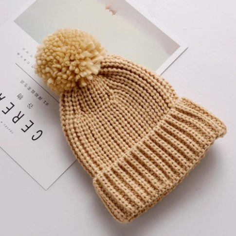 A beige knitted beanie, complete with a cozy faux fur pom-pom atop, from Alaya Junior's "6M-5YRS Kids Winter Hat - Baby & Toddler Girls/Boys" collection rests on a white surface. Its stretchable cotton-acrylic blend promises exceptional warmth for winter adventures. Partially visible beneath the hat is a magazine cover displaying the text "CERE.