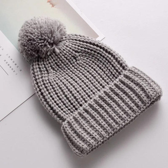 The Alaya Junior 6M-5YRS Kids Winter Hat, a soft knitted cotton acrylic beanie with a faux fur pom-pom, sits on a white surface, perfect for winter adventures for babies and toddlers. Nearby, partially visible pages of a book or magazine add to the scene.