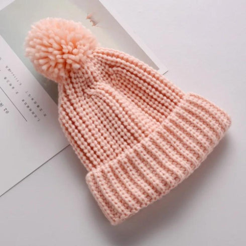 A cozy soft pink beanie from Alaya Junior, featuring a faux fur pom-pom and crafted from stretchable cotton-acrylic fabric, rests on a white surface. Part of a page or document can be seen beneath it, though the text is unclear. This 6M-5YRS Kids Winter Hat is perfect for baby and toddler girls or boys, effortlessly blending style with warmth for winter adventures.