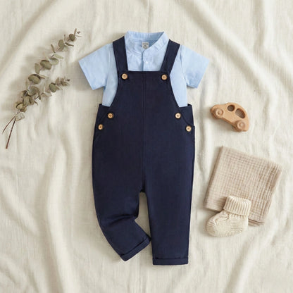 Navy blue overalls with light blue shirt on a beige surface, accompanied by a wooden toy and baby blanket.