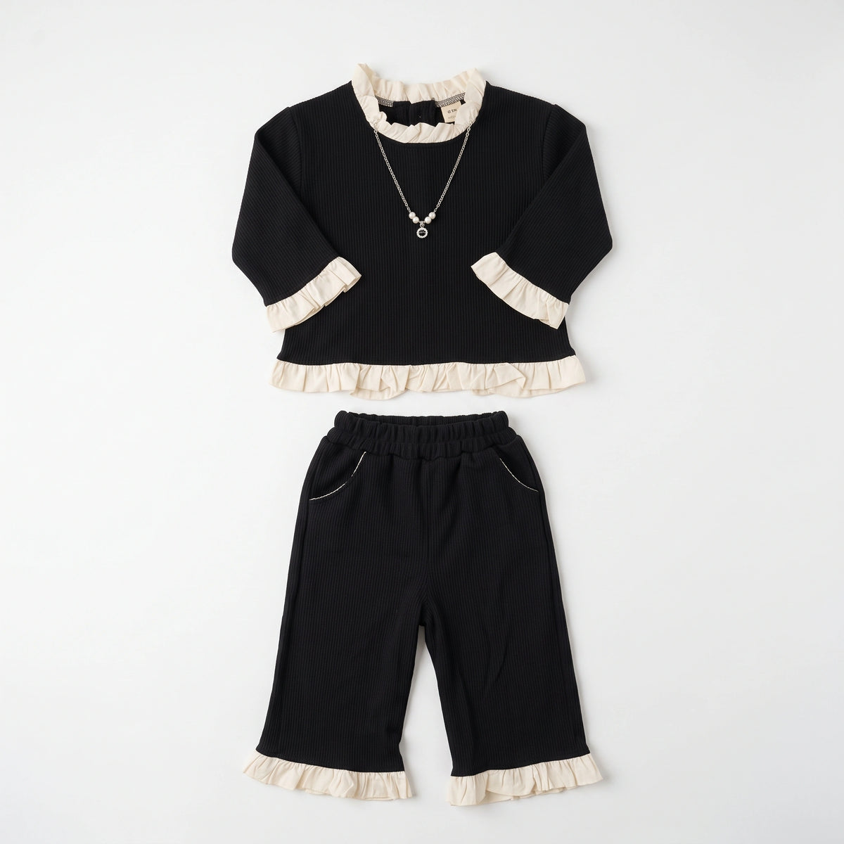A two-piece children's outfit set consisting of a black long-sleeve top with cream ruffle trim at the collar, cuffs, and hem, paired with matching black cropped pants featuring cream ruffle trim at the hem and pocket edges. A silver necklace with a pearl-like pendant is also shown.