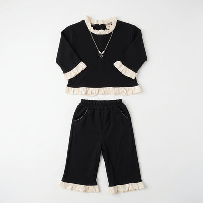 A two-piece children's outfit set consisting of a black long-sleeve top with cream ruffle trim at the collar, cuffs, and hem, paired with matching black cropped pants featuring cream ruffle trim at the hem and pocket edges. A silver necklace with a pearl-like pendant is also shown.