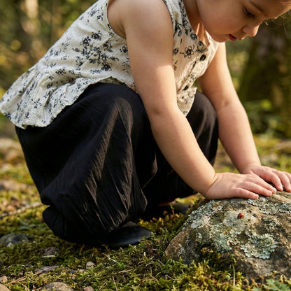 Child sitting on a mossy ground with a rock, wearing a white floral top and dark pants.