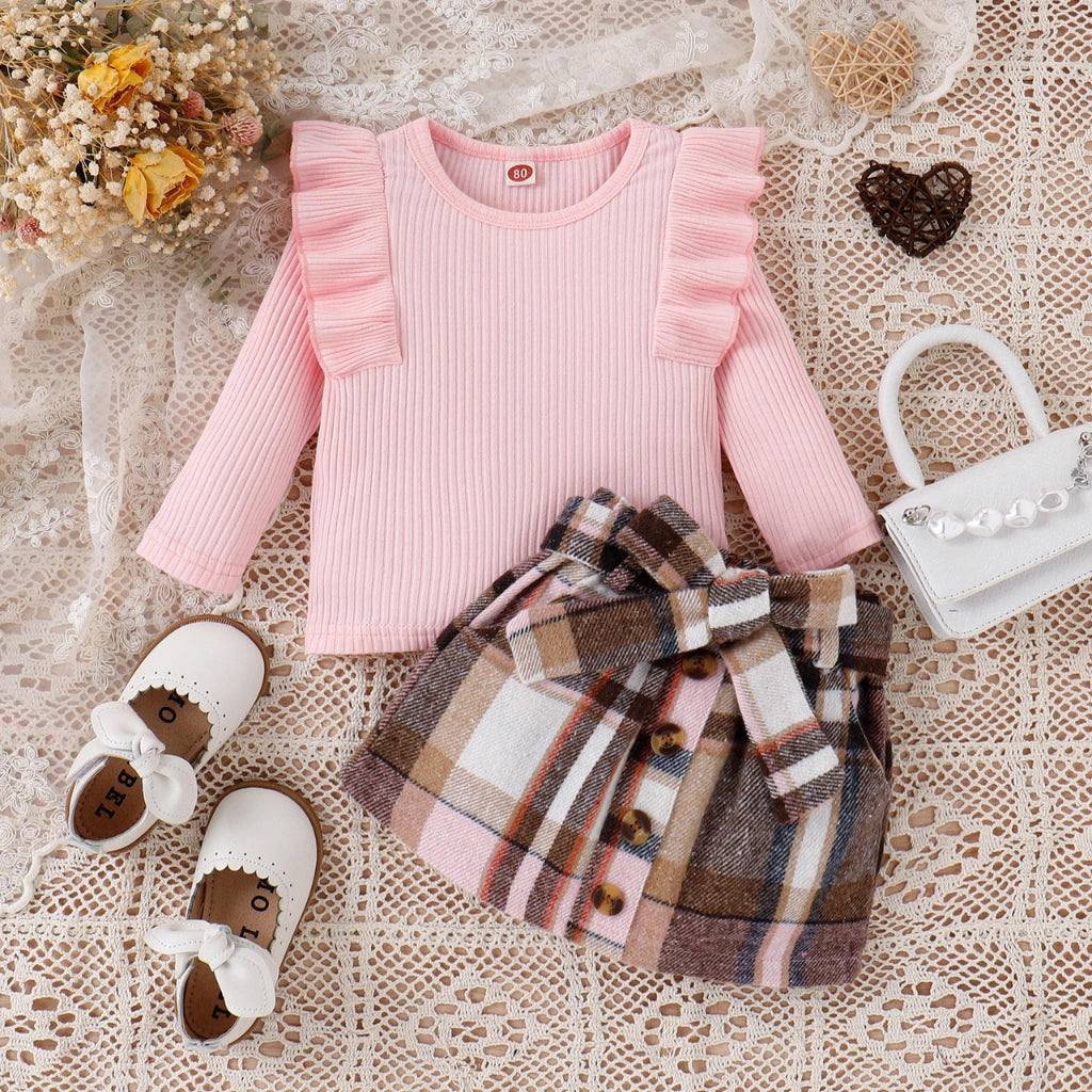 Pink ribbed sweater, plaid skirt, and white shoes on a textured surface with flowers and a heart.