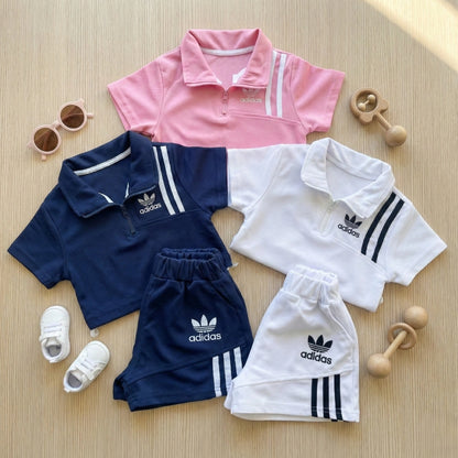 Three children's outfits with Adidas logos on a wooden surface, including a pink polo shirt, navy tracksuit, and white outfit.